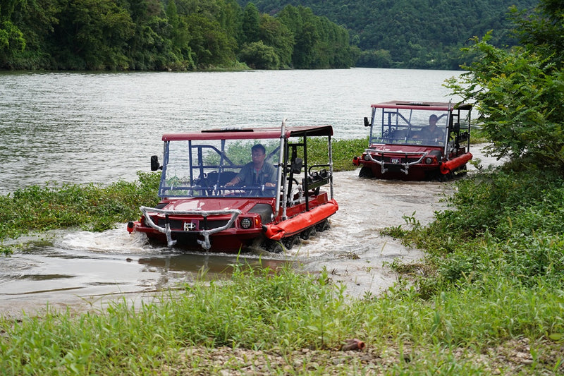 Two red all-terrain vehicles on a muddy path near a body of water with greenery.