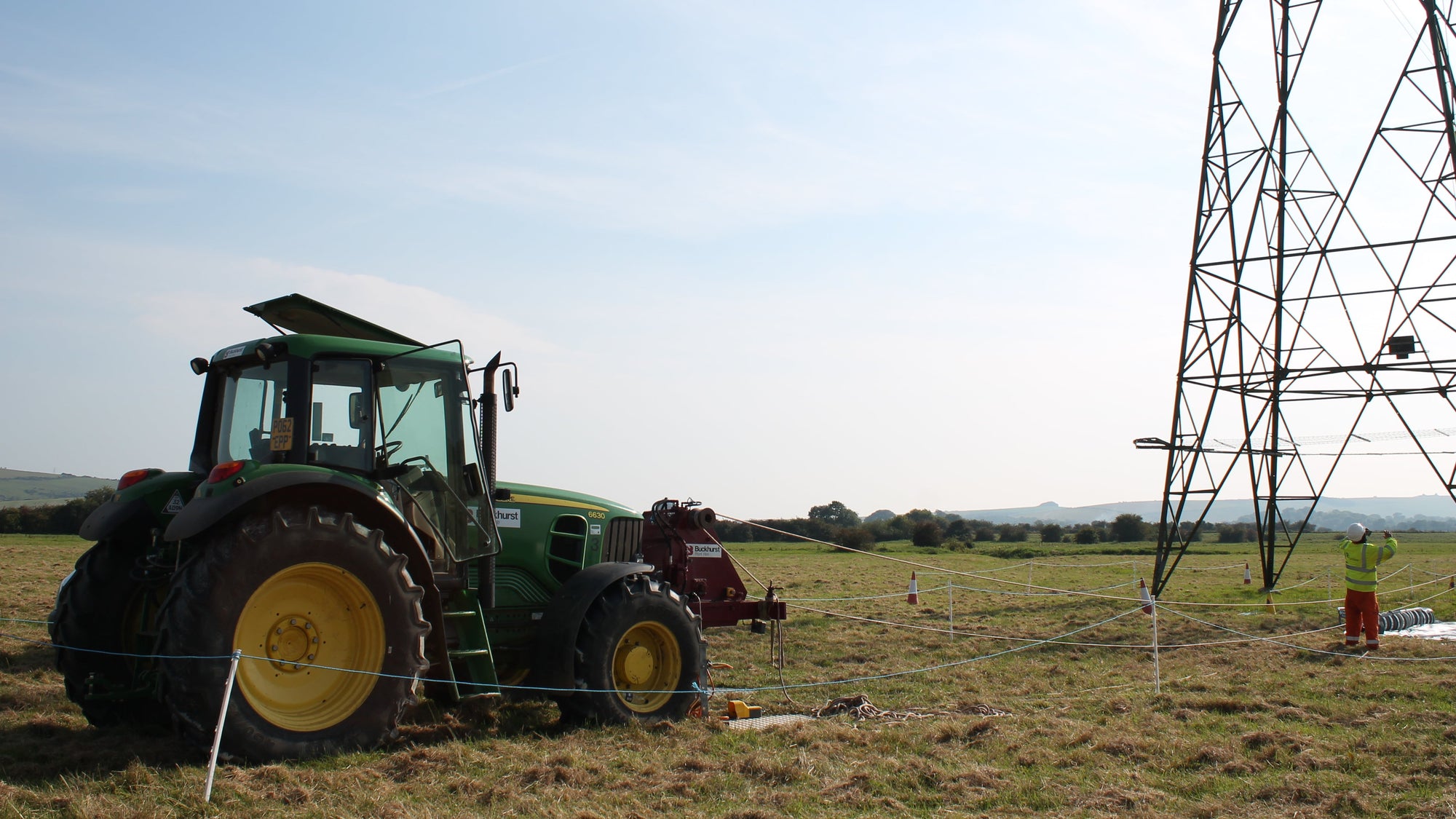 A green John Deere tractor with yellow wheels is parked in a grassy field under a blue sky, with a large metal electricity pylon on the right side of the frame.